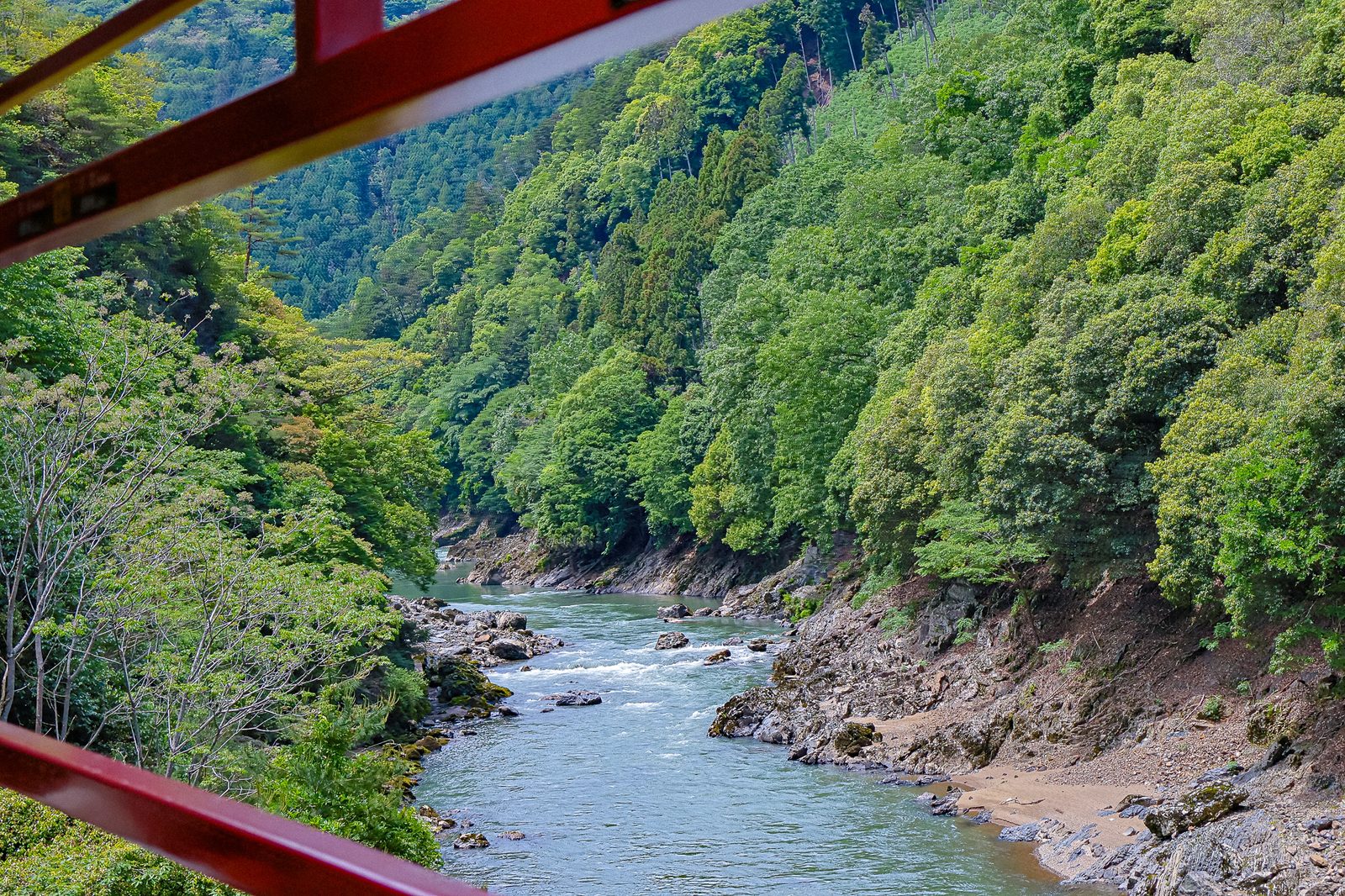 View of the Hozukyo gorge through the red metal framework of Car 5 on the Sagano Scenic Railway