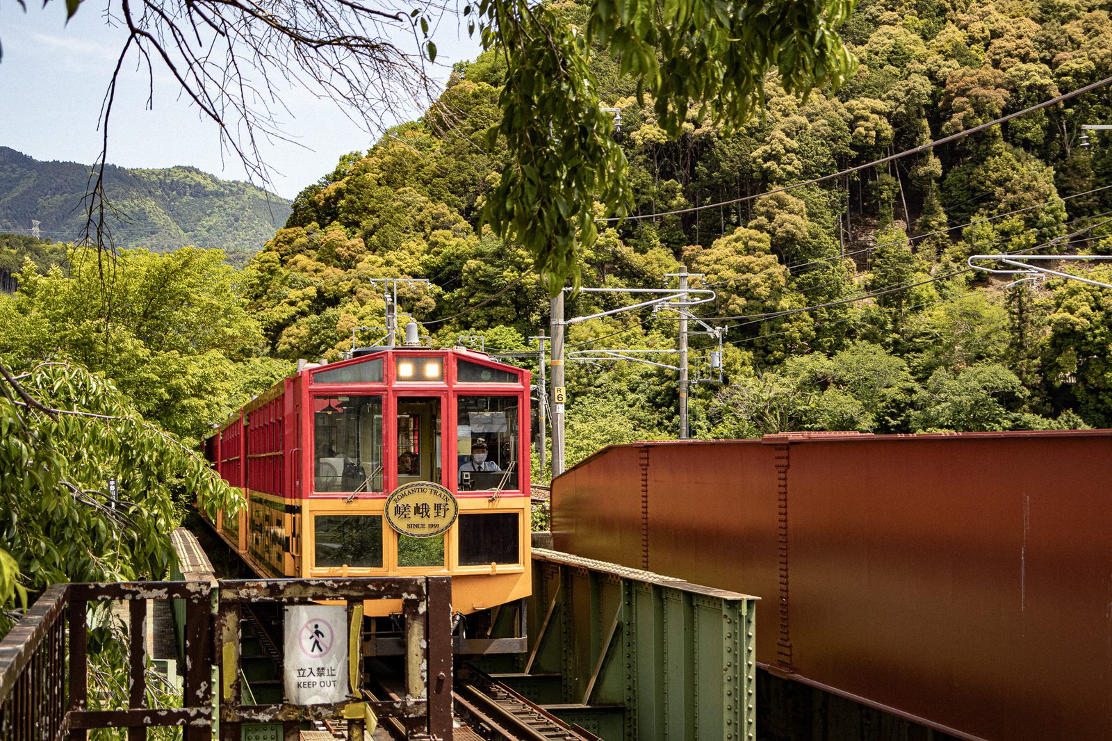 The red and yellow Sagano Scenic Railway train crossing a bridge in the forested Hozukyo gorge